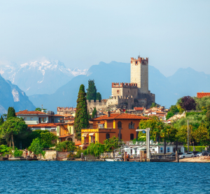 Blick auf das mittelalterliche Castello Scaligero in Malcesine am Gardasee, umgeben von bunten Häusern, mit den Alpen im Hintergrund und blauem Wasser im Vordergrund.