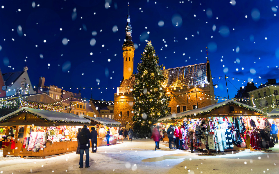 Winterlicher Weihnachtsmarkt in Tallinn mit festlich geschm&uuml;ckten Holzh&uuml;tten, leuchtendem Weihnachtsbaum und Schneeflocken vor der historischen Rathauskulisse.