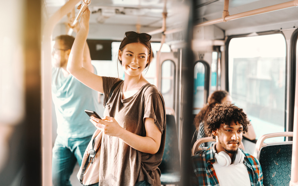 Junge Frau steht in einem &ouml;ffentlichen Bus und h&auml;lt sich fest, w&auml;hrend sie auf ihr Smartphone schaut.