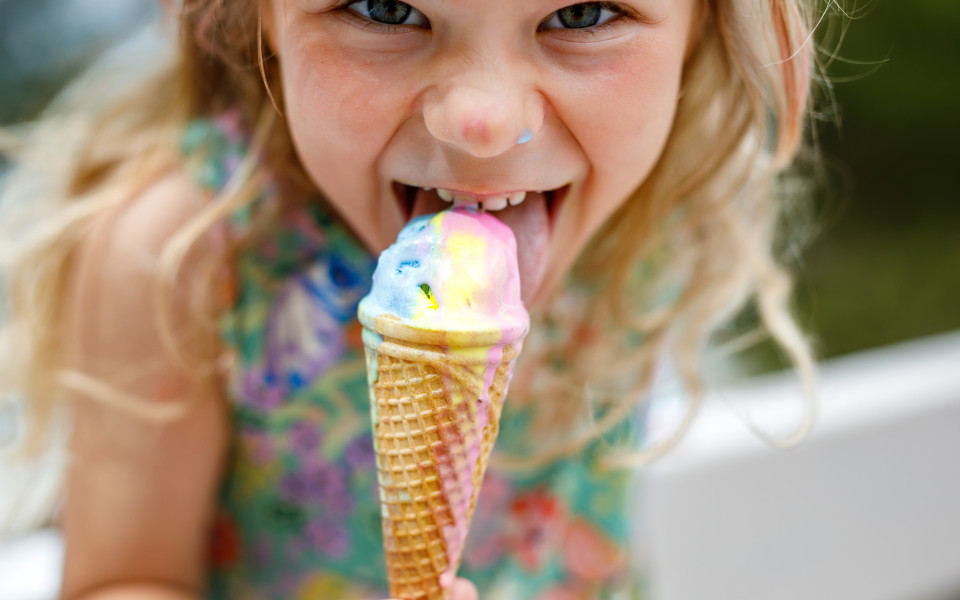 Ein kleines M&auml;dchen mit blonden lockigen Haaren isst ein buntes Eis in einer Waffel. 